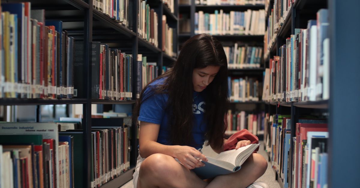 A young woman sitting on the floor of a library, reading a book intently.